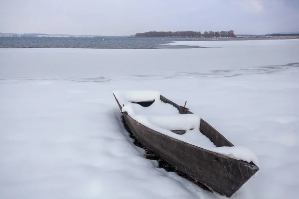 A boat covered with snow on the snowy shore of the lake.