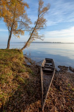 Sonbahar yaprakları arasında, gölün kıyısında bir tekne. Suyun üzerinde eğilen ağaçlar.