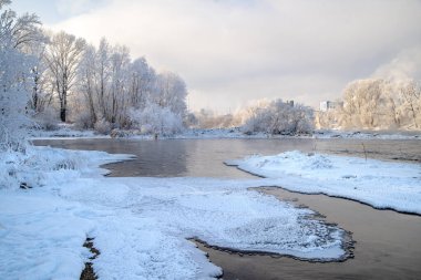 Nehirde kış günü, kısmen buzla kaplı. Sahilde kırağı olan ağaçlar var..