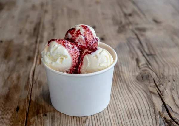 Three balls of white ice cream with berry filling in a cardboard cup on a rough wooden table.