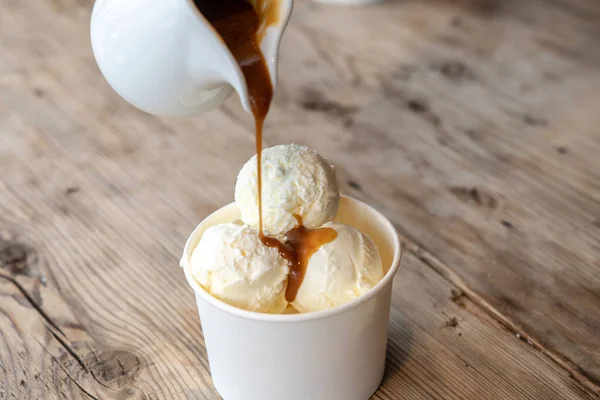 Three scoops of white ice cream are poured over caramel syrup in a cardboard cup on a rough wooden table.
