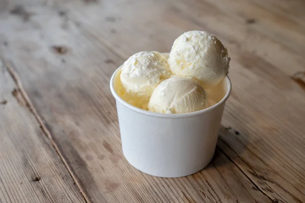 Three balls of white ice cream in a cardboard cup on a rough wooden table.