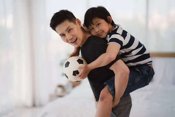 asian dad and son posing with soccer football ball together at home in bedroom. concept of father and son relationship. selective focused