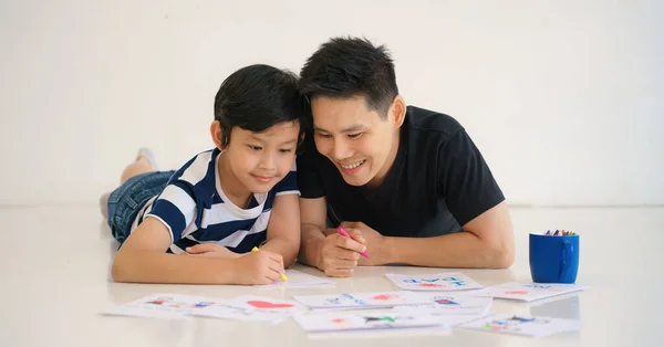 asian dad and son lying on floor together drawing gift card for father day at home. concept of dad and son relationship and father day. selective focused