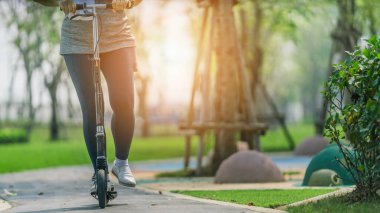Açık havada egzersiz ve arka planda güneş ışığıyla parktaki kaldırımda scooter 'a binen bir kadının egzersizi.