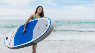 Bikinili güzel Asyalı sörfçü kadın yaz tatilinde denizden sahile yürüyen sörf tahtası taşıyor.