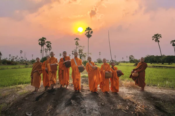 午前中は田の列に並んでお椀を持つ僧侶が先頭に立つ仏教徒の初心者で 人々から食べ物を受け取るためにタイの田舎の村への道に沿って太陽が昇ると — ストック写真