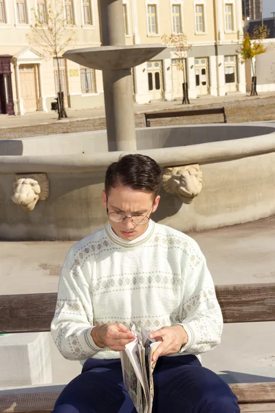 Man with glasses in white sweater reading newspaper on bench — Stock Photo, Image