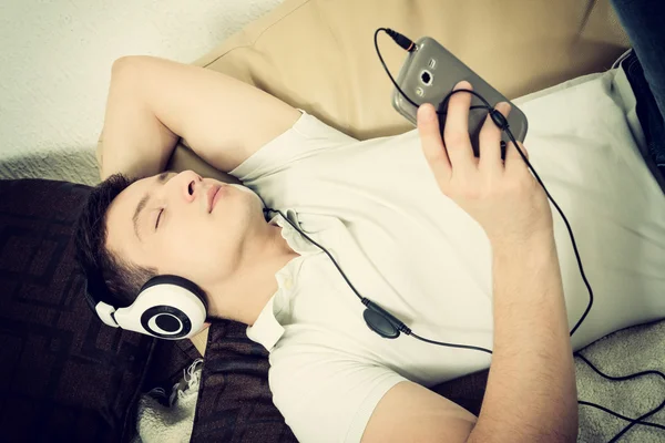 Hombre en el sofá sosteniendo teléfono inteligente escuchando música con auriculares —  Fotos de Stock