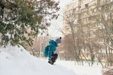 Tepedeki yeşil ceketli ve şapkalı çocuk. Çocuklar karda oynar. Kış şehri.