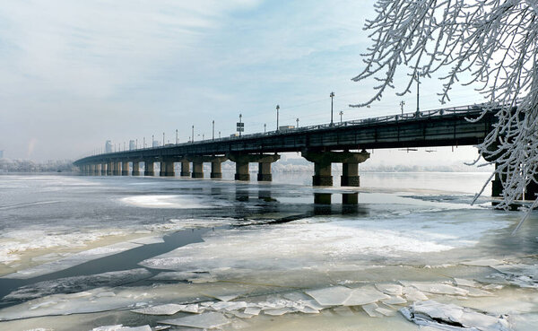 Paton Bridge over a frozen river in winter. Icy water, cracked ice, and frosty trees create a cold urban landscape with a famous city bridge under a pale winter sky.