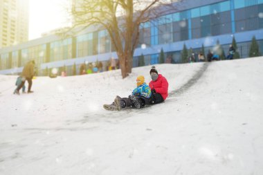 Ebeveyn ve çocuk kış parkındaki karlı yamaçta birlikte kayıyorlar, aile açık havada eğlence, soğuk hava aktivitesi, neşeli bir çocukluk tatili.