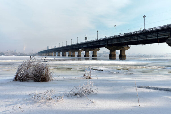 Paton Bridge over a frozen river in winter. Icy water, cracked ice, and frosty trees create a cold urban landscape with a famous city bridge under a pale winter sky.