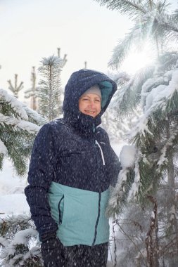 Sıcak kış ceketi giymiş gülümseyen bir kadın kar altında çam ağaçlarının arasında duruyor açık havada, açık havada, açık bir ormanda, aydınlık bir kış gününde..