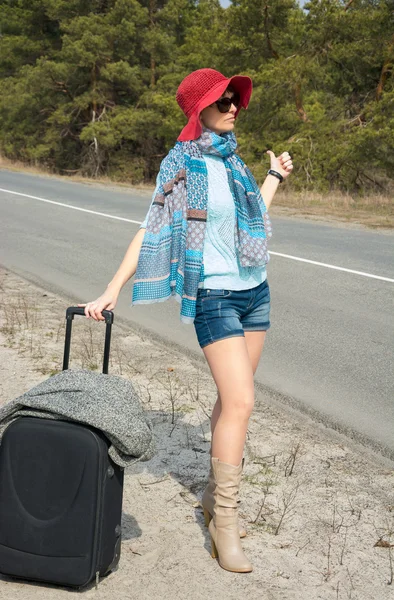 Jeune femme avec une valise est auto-stop sur la route pointant Image En Vente