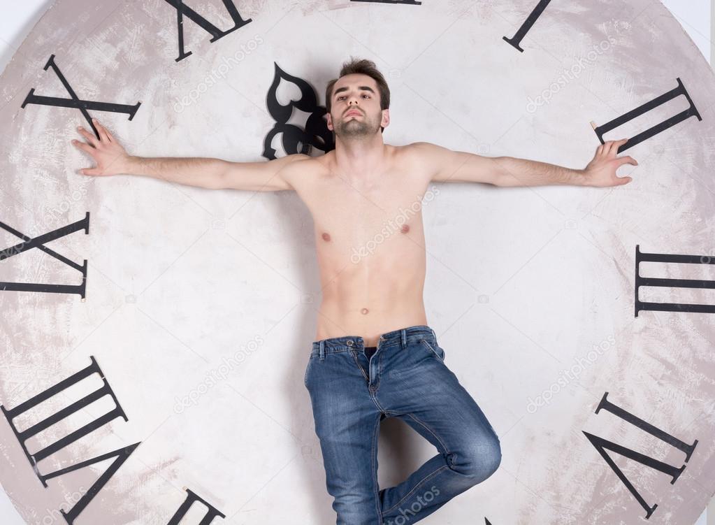 A shirtless young man is standing in front of a clock