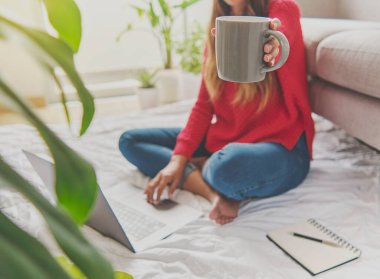 Çay içen, bir apartmanın zemininde oturan, notlar yazan bir kadın, önünde bir dizüstü bilgisayar açtı. Merkez ofis konsepti. Kahve ikram ediyorum. Evde yaratıcı iş kavramı. Seçici odak.