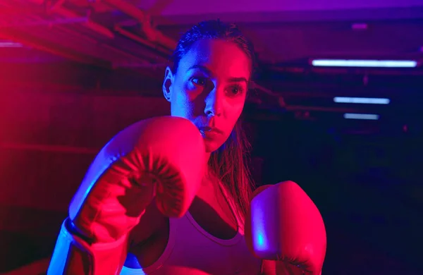 Beautiful female boxer training, red boxing gloves, ring with red and blue lights, industrial gym, neon lights. selective focus
