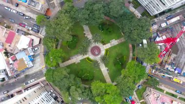 İnsansız hava aracı görüntüsü yemyeşil yollarla ve çevre şehir sokaklarıyla dairesel bir şehir parkının üzerinde uçuyor.