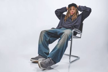 Sandalyesinde oturan, sokak kıyafetlerini sergileyen ve günlük kıyafetleri olan genç bir adam.