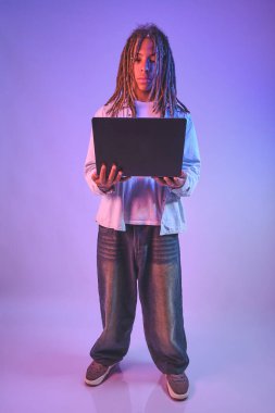 Rastalı ergen ayakta duruyor ve neon ışık yansımalı bir dizüstü bilgisayara konsantre oluyor.