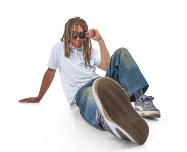 Rastalı ve güneş gözlüklü genç bir adam beyaz bir arka planda günlük giysiler içinde oturuyor.