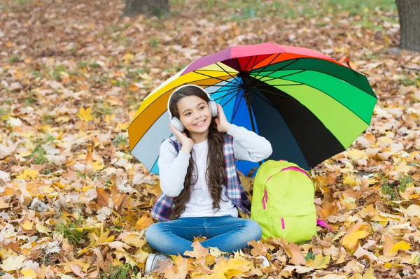 Vidám tini lány pihenni őszi erdőben vagy parkban, miközben zenét hallgatni fülhallgató közelében iskolai hátizsák alatt színes ernyő, online oktatás — Stock Fotó