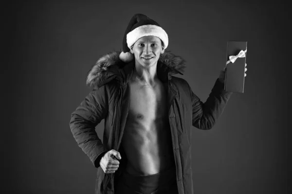 愛の贈り物だ。大人のための新年会。サンタ・ハットのマチョ・マンとパフ・ジャケット。セクシーなサンタ・クラスのレッド・バックグランド。筋肉質の男プレゼントボックス。クリスマスだ。クリスマスの冬休み。外は寒くて — ストック写真