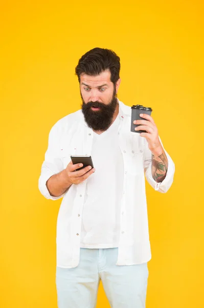 Chico sorprendido con bigote y barba beber café y leer chat en el teléfono inteligente. hombre barbudo maduro comunicarse por teléfono mientras bebe café de la mañana. bebida caliente en taza de papel. la vida moderna — Foto de Stock
