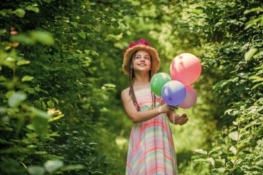 Güzelliğin kaynağı. Parti balonlarıyla güzel bir kız. Küçük kızın şapkasında çiçek açan güller var. çiçek kompozisyonu. Hasır şapkalı güzel kız balonları tutuyor. Mutlu çocukluk