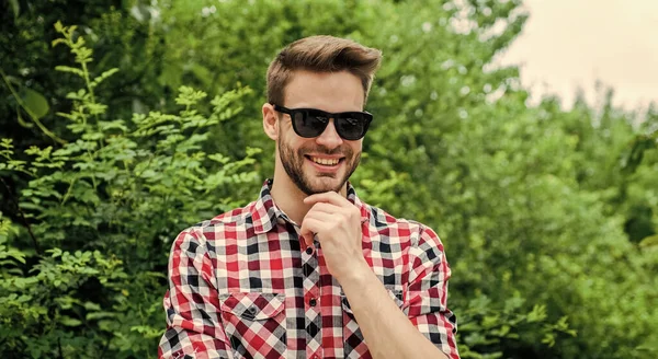그의 스타일에 대한 확신. 웃는 남자는 체크무늬 셔츠와 선글라스를 착용 한다. 여름 방학 개념이야. 화려 한 야외 생활을 하는 잘생긴 남자. 남성 패션과 아름다움. 섹시 한 수염의 마초. 평상복 — 스톡 사진