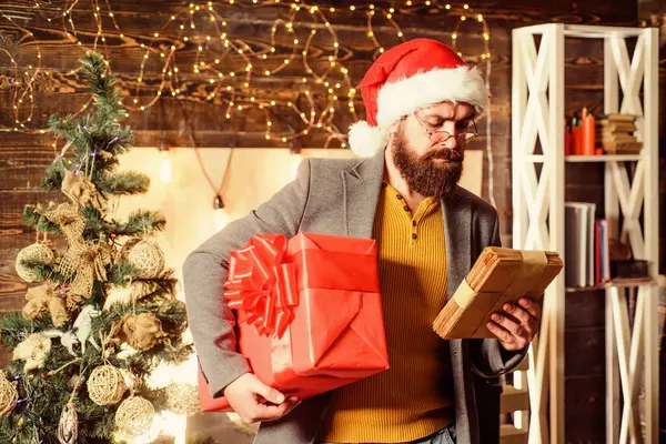 Julstämningen är här. Skäggig kille bär tomten hatt bära julklapp låda. Leverans julklapp. Julklapp. Man tomte hatt hålla gåva. Leveransservice. Sprid lycka och glädje — Stockfoto