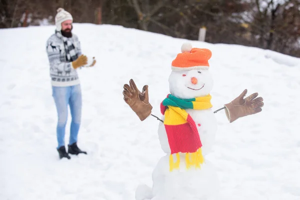 Snowman and cheerful bearded hipster knitted hat and warm gloves play with snow outdoors. Have fun winter day. Let it snow. Christmas holidays. Active lifestyle. Snow games. Leisure on fresh air