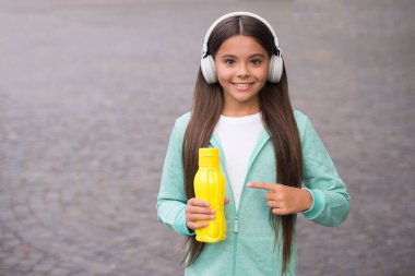 Yüzünde kulaklıkla gülümseyen liseli kız plastik şişeyi işaret ediyor açık havada su içiyor, fotokopi çekiyor, susamış.