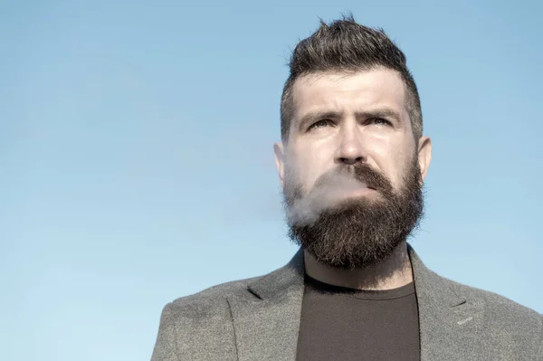 Homem com barba e bigode fumando fundo céu azul. Tabagismo e hábitos conceito. Hipster com cabelo elegante rosto concentrado pensativo com fumaça branca voando para fora da boca. Fumar ao ar livre — Fotografia de Stock