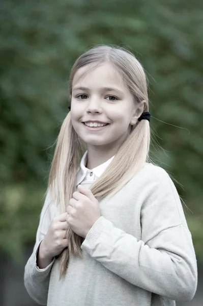 Penteado e conceito de cuidado do cabelo. Menina no rosto sorridente posando com duas caudas, fundo natural verde. Menina gosta de olhar bonito, elegante e elegante. Menina criança com cabelos longos parece adorável — Fotografia de Stock