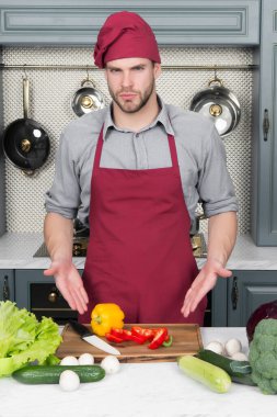 Yemek pişirmek için gerekli malzemeler. Adam masadaki sebzeleri işaret ediyor. Mutfakta aşçı şapkası ve önlüğü var. Vejetaryen menüsü ve sağlıklı beslenme. Yemek hazırlama ve yemek tarifleri