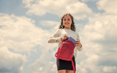 Güneşli bir günde küçük bir çocuk. Yaz tatili. Küçük çocuk bahar havasında gülümsüyor. Kıvırcık kız sırt çantasını gökyüzünde tutuyor. Okula dönüyorum. çocukluk mutluluğu. Çalışmaya odaklan.