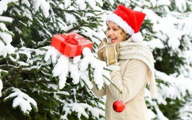 Ziller çalıyor. Satış ve indirim. Mutlu kadının elinde Noel hediyesi kutusu var. Noel ağacı süslemeli kız. Bayan Kırmızı Noel Baba şapkası kürk ağacında. Kış alışveriş mutluluğu. Açık hava mükemmel bir ruh halidir.