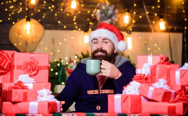 Hipster prepared gifts for family. Generous new year. Lot of gifts. Wrapped gifts with ribbons and bows. Happy winter holidays. Warming up. Tea time. Man bearded santa claus hat drinking hot beverage