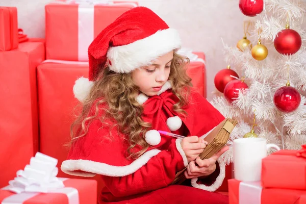 Wenslijst. Kind santa kostuum genieten van kerstavond. Kind schrijft brief aan de kerstman. Lieve kerstman. Meisje houdt pen en papier in de buurt van kerstboom en geschenkdoos. Geloof in wonderen. Stuur brief voor de kerstman — Stockfoto
