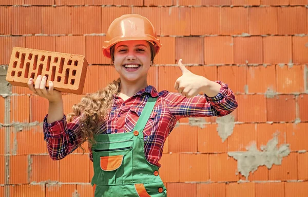 Little worker in helmet building from brick. Architecture construction child. Child in uniform working around brick wall. labour day concept. Little girl bricklayer. learning how to build. thumb up