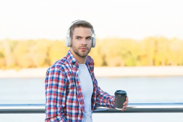 bearded man listen music in digital earphones and drinking coffee to go from paper cup wearing checkered shirt outdoor, refreshment