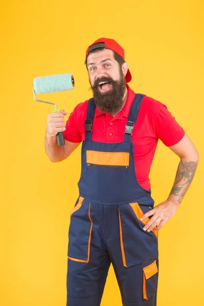 Trabalhador pintor masculino barbudo surpreso em uniforme de terno de caldeira usando ferramenta de rolo de pintura para pintura de parede em cor amarela, construindo — Fotografia de Stock