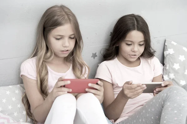 Children in pajama interact with smartphones. Application for kids fun. Internet surfing and absence parental advisory. Smartphone internet access. Girls sisters wear pajama busy with smartphones