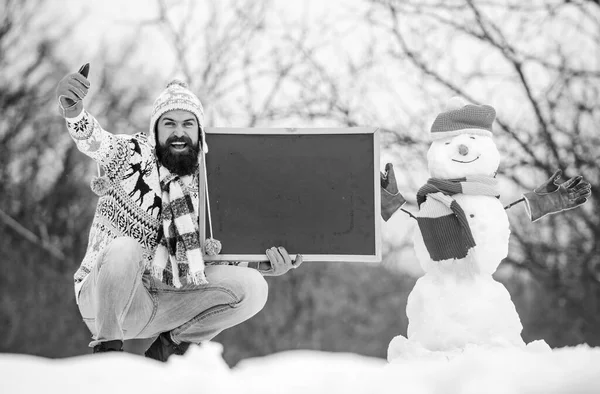 Winter announcement. Hipster knitted hat and gloves show blackboard. Winter holidays. Guy and snowman snowy nature background. Winter event. Upcoming event. Man with beard hold chalkboard copy space — Stock Photo, Image