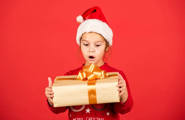 ¡Increíble! Increíble sorpresa. Feliz Navidad y felices fiestas. Vacaciones de invierno. Compras de regalos de Navidad. Paquete regalo. Pequeña adorable niña Santa sombrero celebrar caja de regalo. Niño mantenga la caja actual fondo rojo —  Fotos de Stock