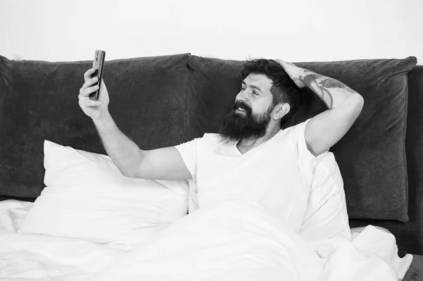 早上第一件事就是自私自利。戴胡子的男人在床上自私自利.快乐的嬉皮士微笑着对着相机手机.视频自拍。博客的自拍。社交网络。网络博客。现代生活。新技术 — 图库照片