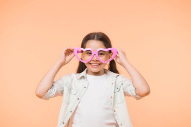Mutlu çocuk sarı arka planda kalp şeklinde gözlük takıyor. Moda aksesuarlarıyla gülümseyen küçük kız. Yaz modası görünüşü. Funk stili güzellik. Genç ve kendinden emin. Sevgililer Günü. Seni seviyorum.