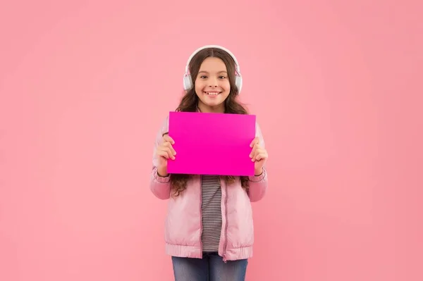 Criança alegre usando fones de ouvido e mostrando papel vazio para espaço de cópia, anúncio — Fotografia de Stock